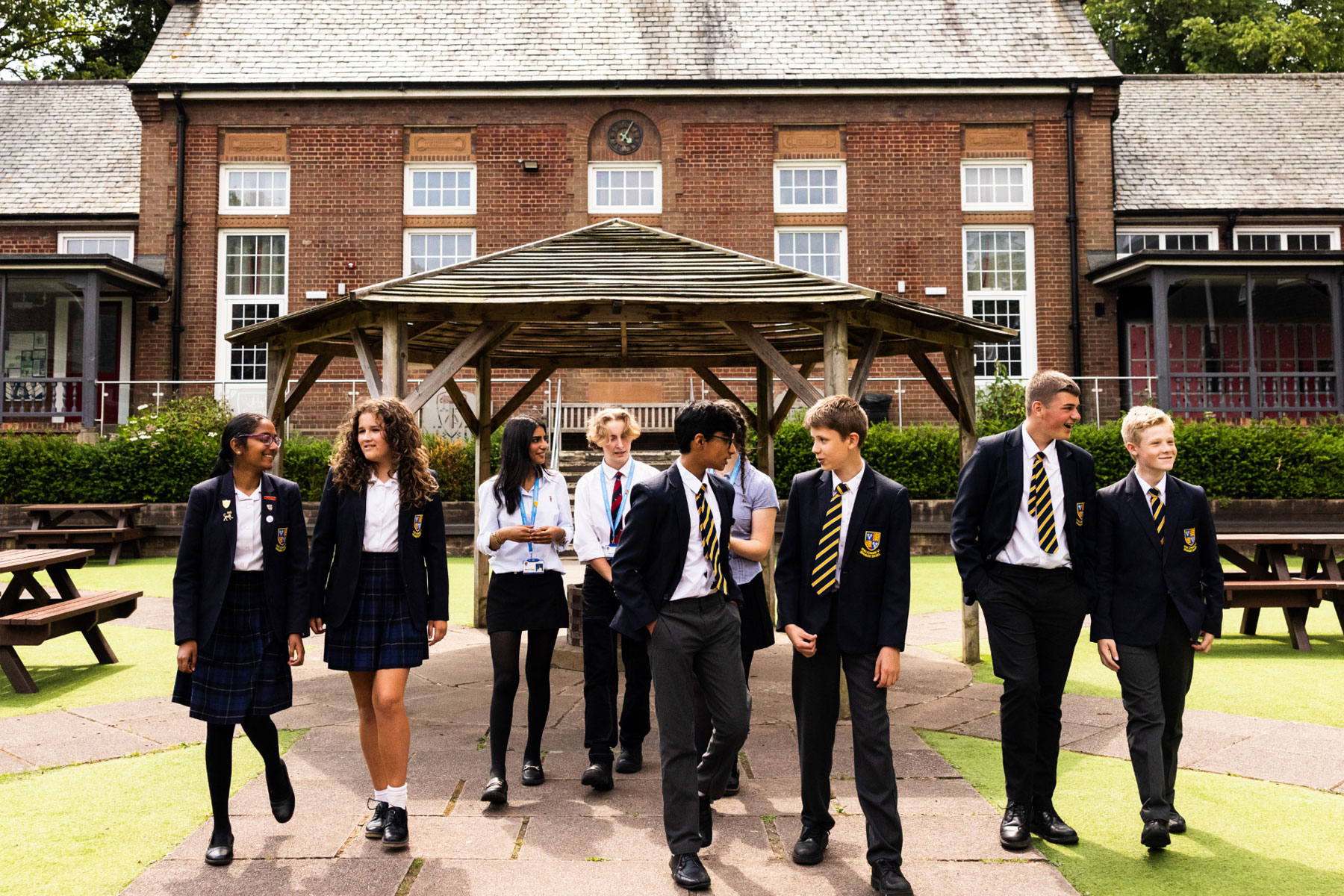 Students in school uniforms walking together outside the main entrance of King Edward VI Grammar School, engaged in friendly conversation. The historic school building with large windows and greenery surrounds them, creating a welcoming and vibrant atmosphere. No visible text in the image.